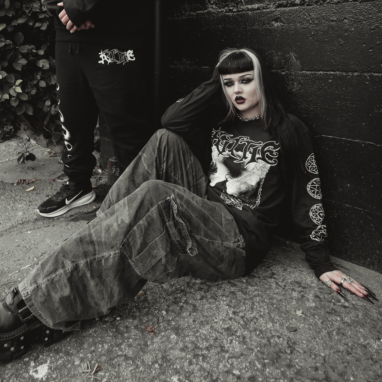 A person with black and white hair, dramatic makeup, and dark clothing—including the Kittie Eclipse Black Long Sleeve—sits against a black wall. Another figure in black stands partially visible, enhancing the goth vibe.