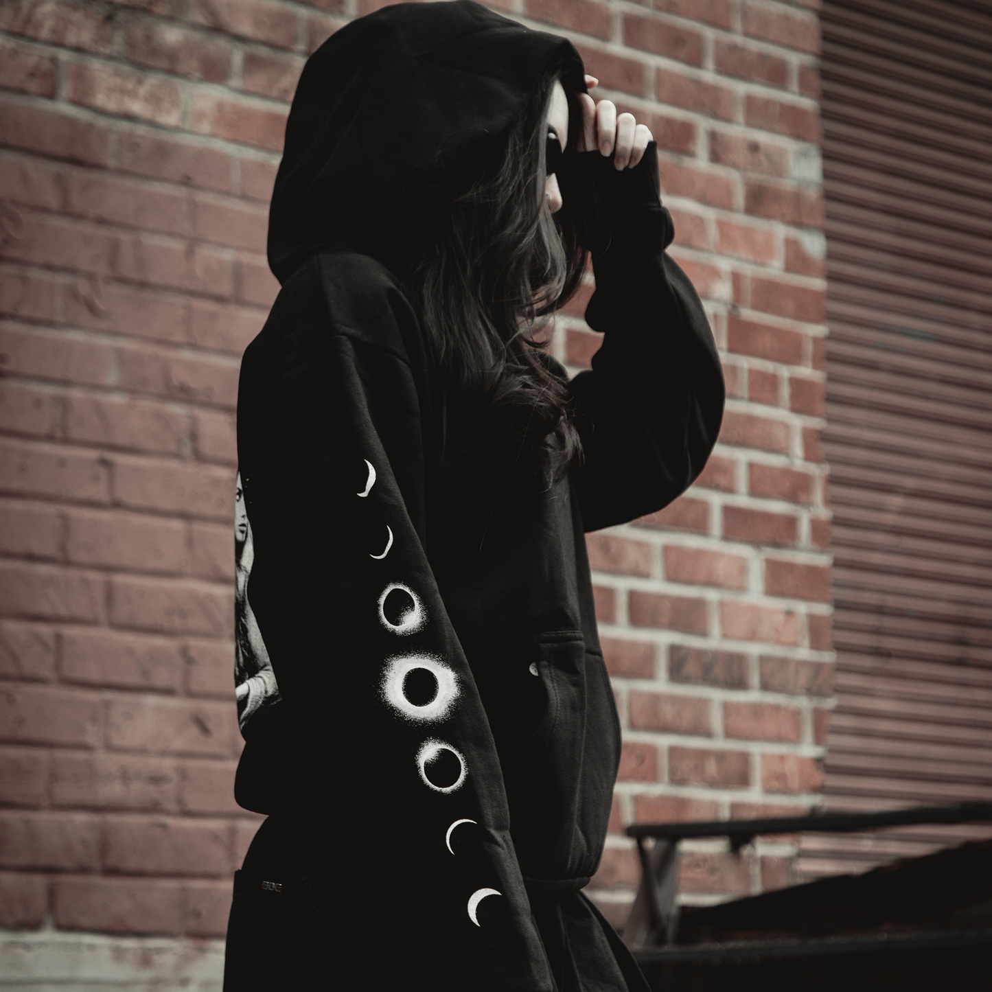 A person in a Kittie Hecate - Black Pullover, featuring moon phase sleeve designs, stands against a red brick wall with the hood partly concealing their face. The oversized, heavyweight fleece hoodie combines comfort and style.