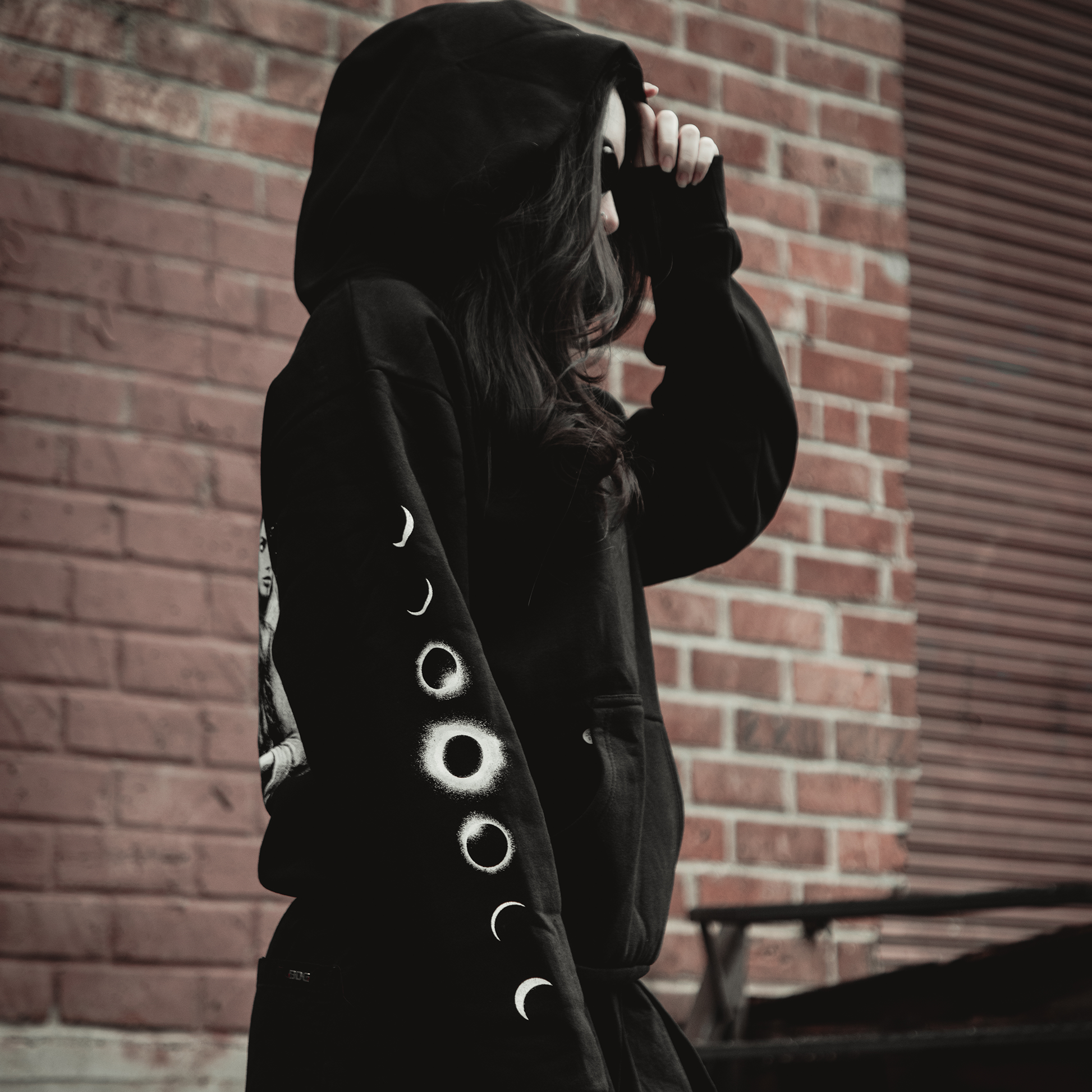A person in a Kittie Hecate - Black Pullover, featuring moon phase sleeve designs, stands against a red brick wall with the hood partly concealing their face. The oversized, heavyweight fleece hoodie combines comfort and style.