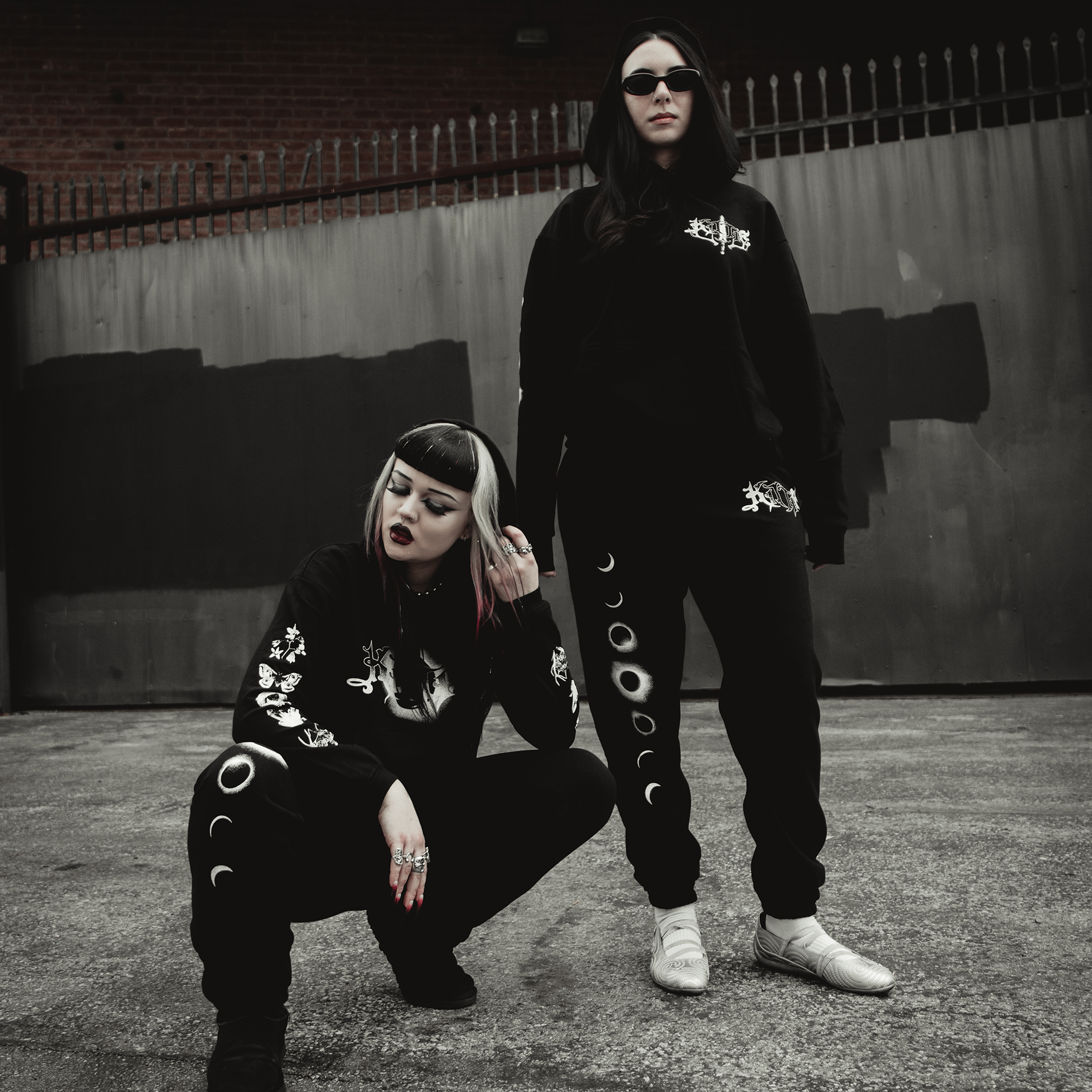 Two women in black streetwear pose confidently by a metal fence and brick wall, wearing Kittie's Hecate - Black Sweatpants with moon designs. One crouches, the other stands; both sport dark sunglasses and bold makeup.