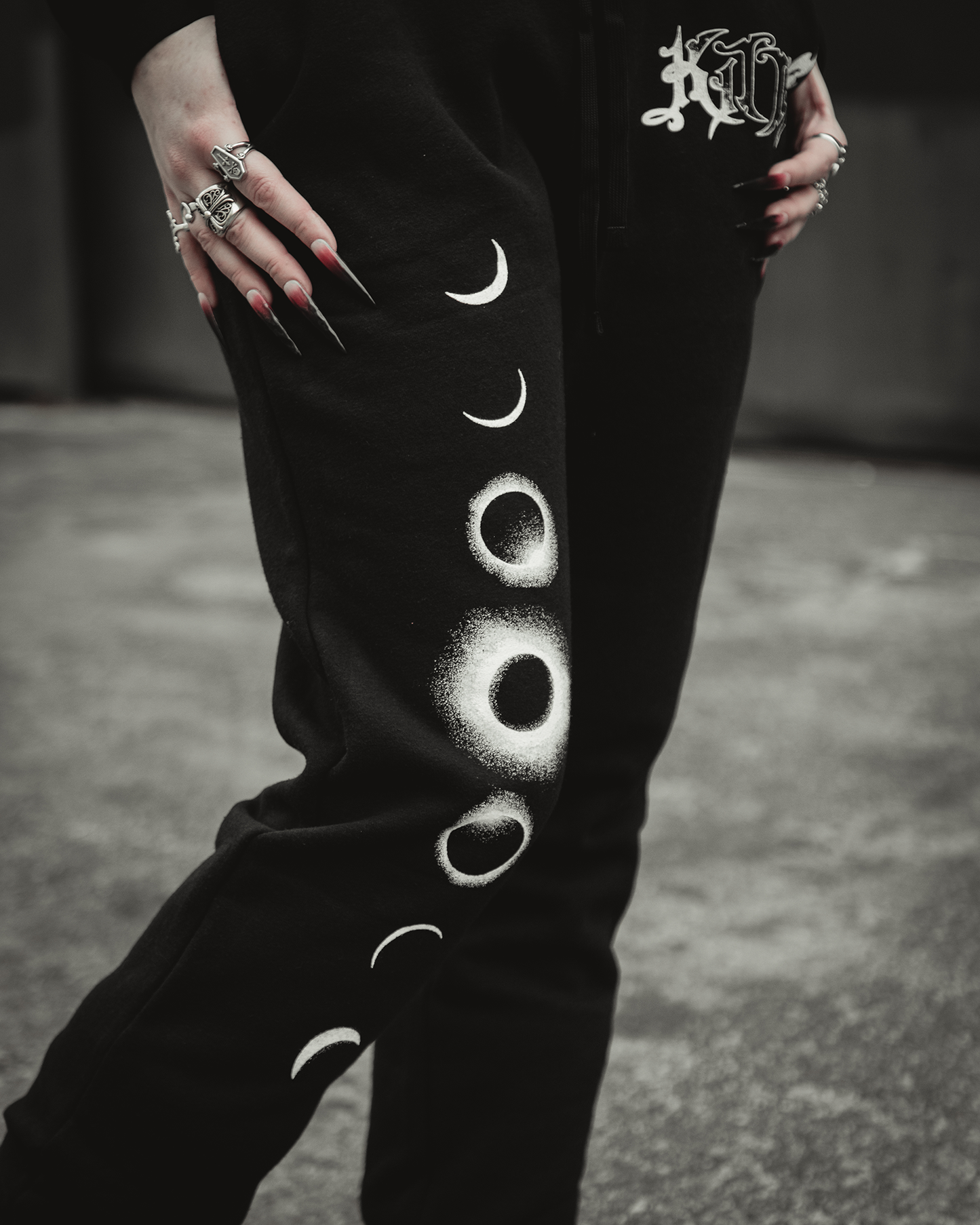 A person stands on gray pavement wearing Kittie’s Hecate - Black Sweatpants, featuring white moon phases down the leg. They accessorize with silver rings and long red nails, adding an edgy touch to their relaxed style.