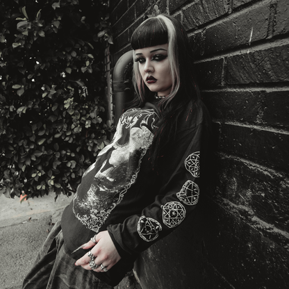 A young woman with dramatic makeup and two-toned hair leans against a brick wall, wearing the Kittie Eclipse - Black Long Sleeve shirt with occult designs, standing on a sidewalk beside leafy bushes.
