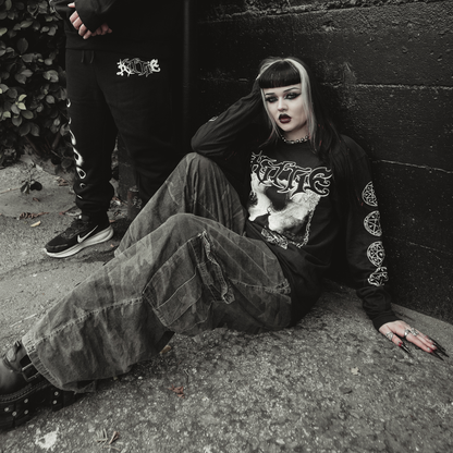 A person with black and white hair, dramatic makeup, and dark clothing—including the Kittie Eclipse Black Long Sleeve—sits against a black wall. Another figure in black stands partially visible, enhancing the goth vibe.
