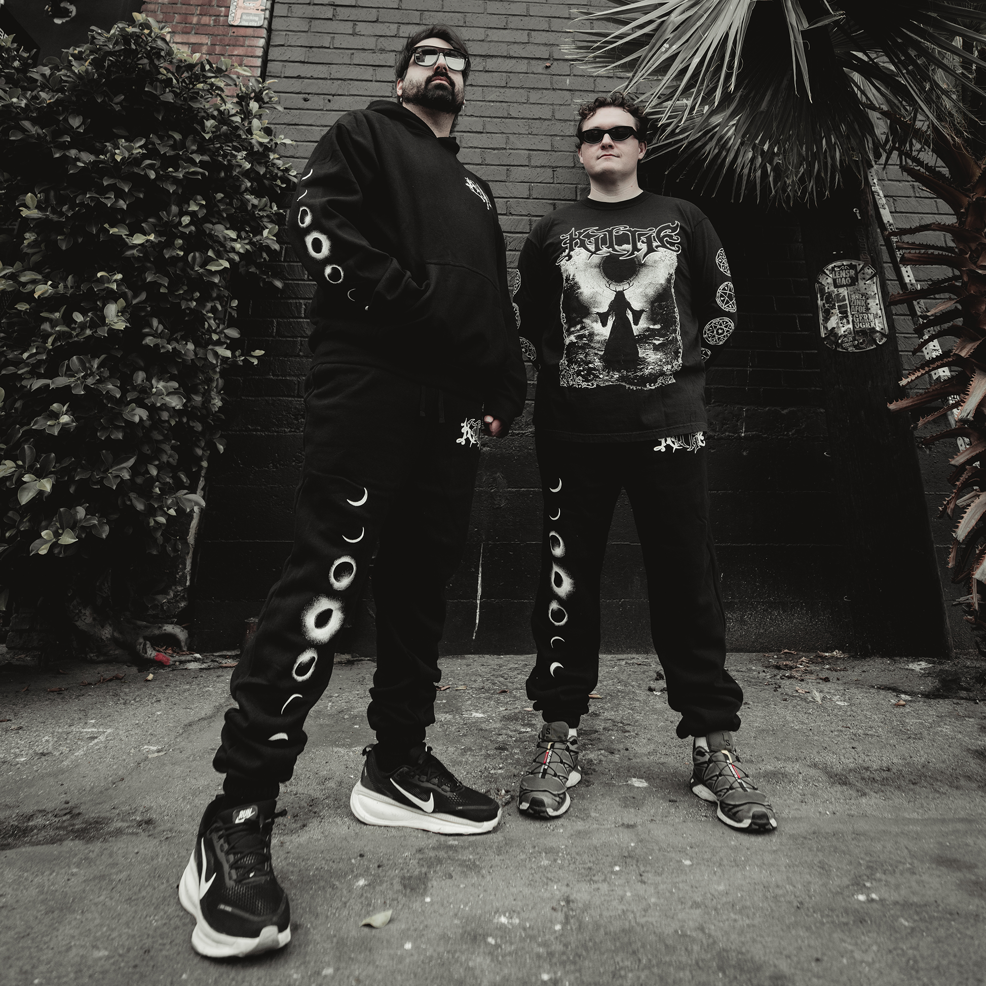 Two people in black streetwear with moon phase and occult graphics stand on a city sidewalk with urban plants and brick wall behind them. Their relaxed-fit outfits feature Kittie's Hecate - Black Sweatpants.