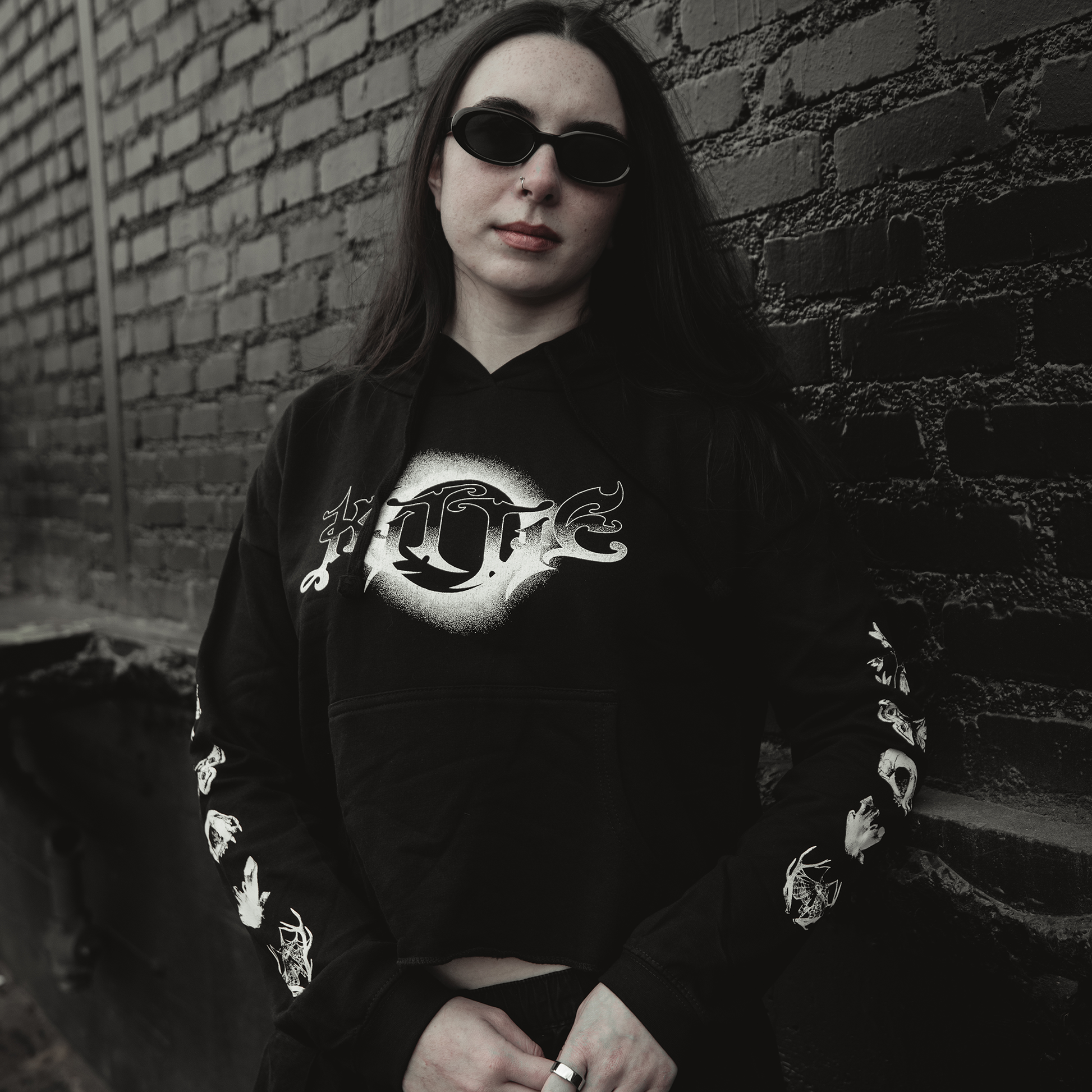 A woman with long dark hair and black sunglasses wears the Kittie Nox - Black Cropped Women's Pullover, featuring white graphic designs, while standing against a dark brick wall with a serious expression.